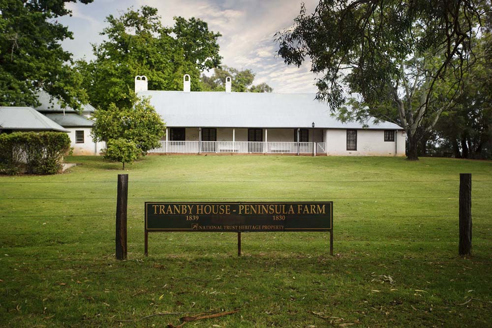 Historic Tranby House-Peninsula Farm (1839-1830), National Trust heritage property in Maylands: white colonial building with tin roof, verandas, surrounded by lush green lawns, trees and cloudy sky. Timeless charm in premium Perth real estate by trusted real estate agents.